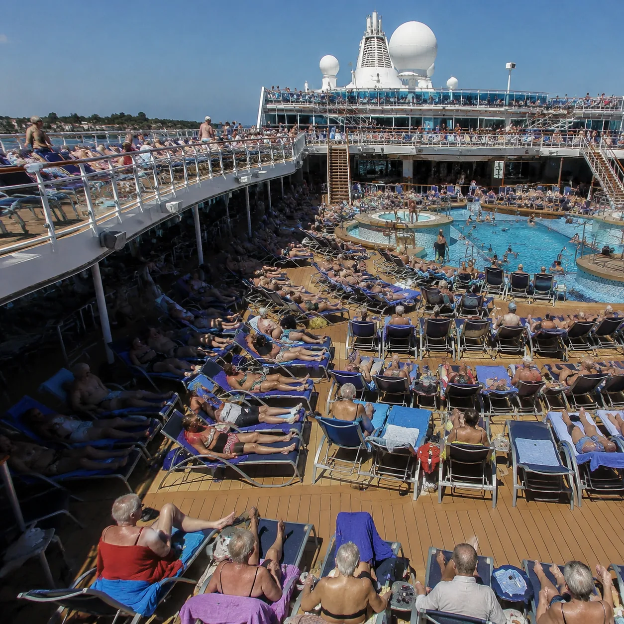 Realistic editorial photo of cruise pool deck game with passengers laughing, towels, and lounge chairs nearby, sunny ene