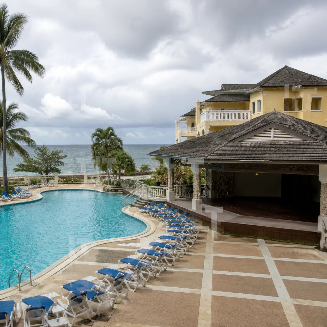 Square photo of an adults-only Jamaica resort pool deck with quiet but dated tropical buildings, empty stage area, cloud