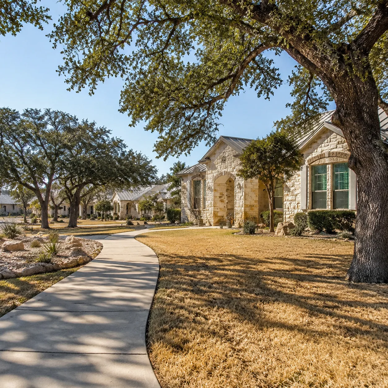Realistic editorial photo of a San Antonio active adult community with stone-front homes, dry grass, live oaks, and hot 