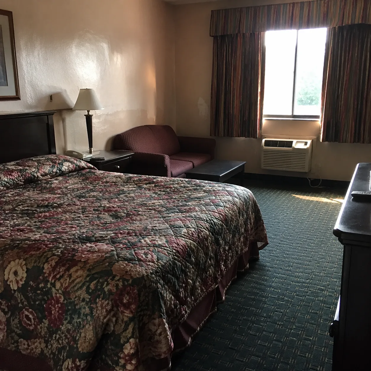 Mid-budget hotel room with dated floral bedspread, scuffed walls, an old air conditioning unit under the window, harsh m