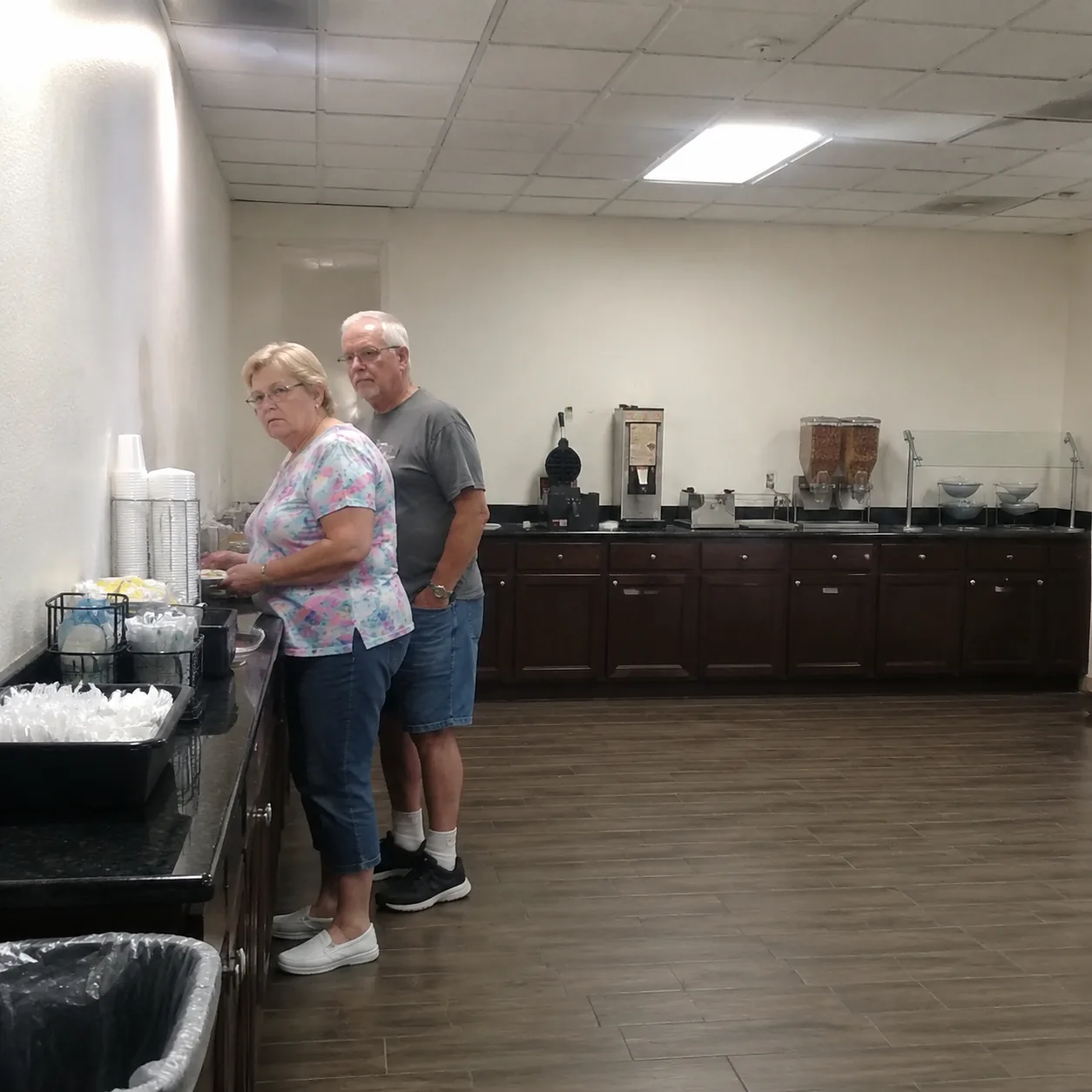 Hotel breakfast area with nearly empty buffet, single waffle machine, sparse cereal dispensers, styrofoam plates and pla