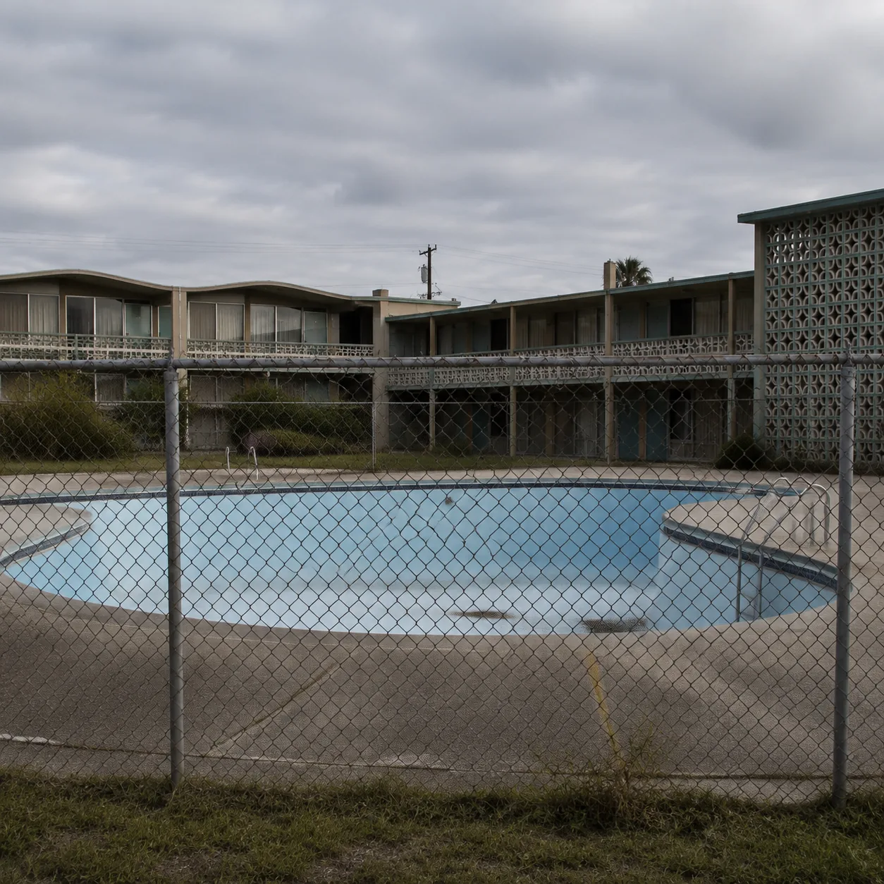 Aging mid-century roadside hotel with empty swimming pool behind chain link fence, cracked concrete pool deck, vintage 1