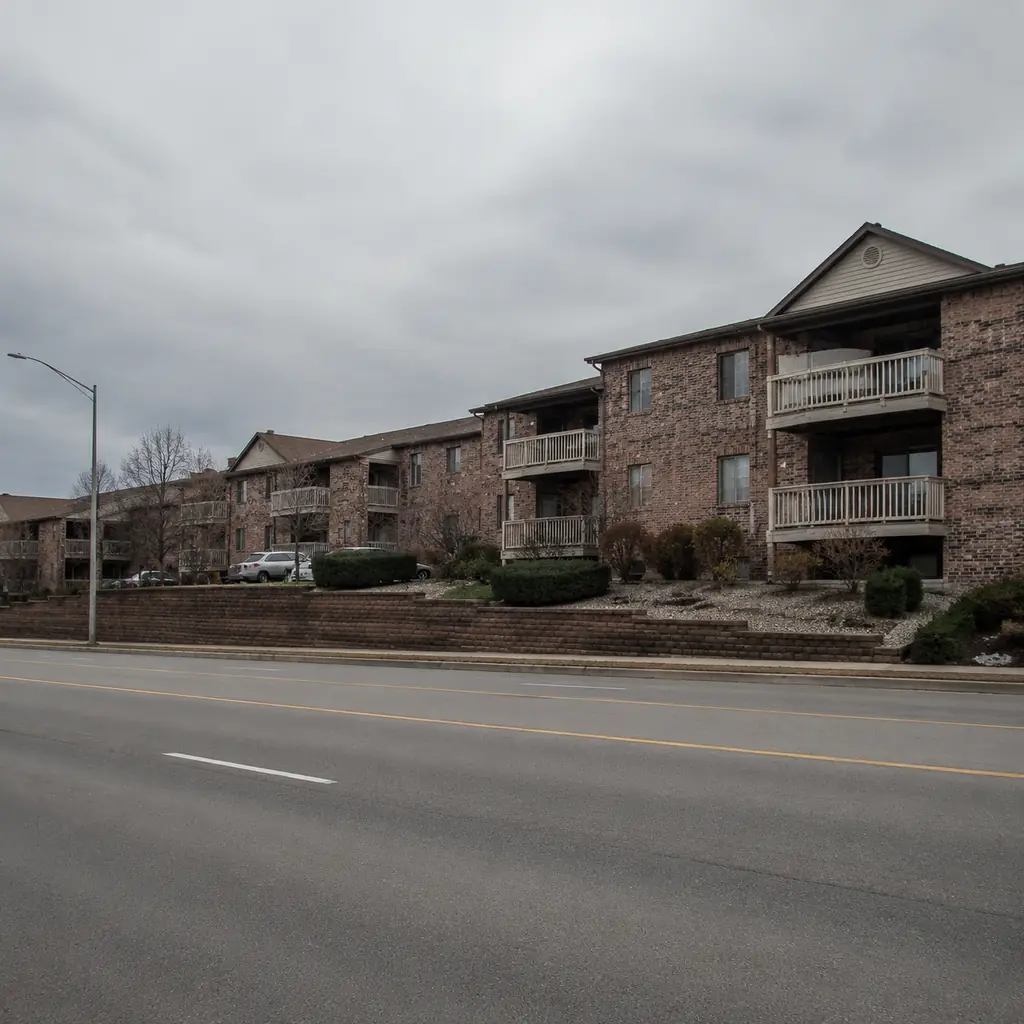Realistic editorial photo of a modest Ohio retirement community on a busy suburban road with brick buildings, small balc