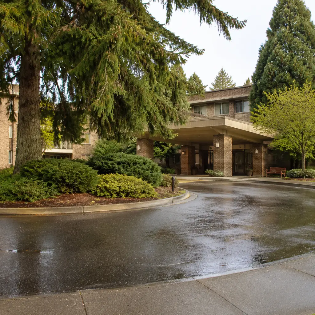 Realistic editorial photo of a 1980s era brick retirement campus with a circular drive, covered entry and mature evergre