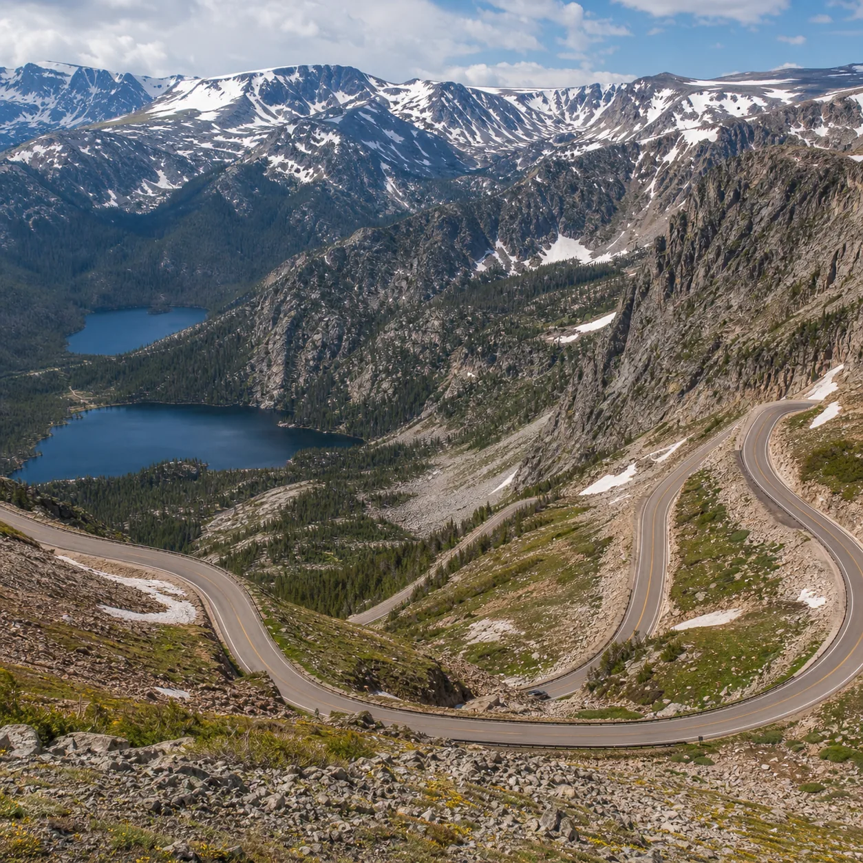 Realistic editorial photo of a high alpine highway near Beartooth Pass with switchbacks, mountain lakes, and patches of