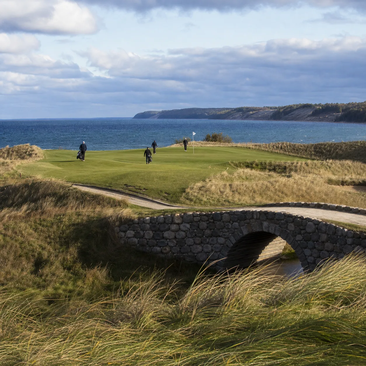 Realistic editorial photo of a rugged Lake Michigan links-style golf course with fescue dunes, blue lake water, stone br