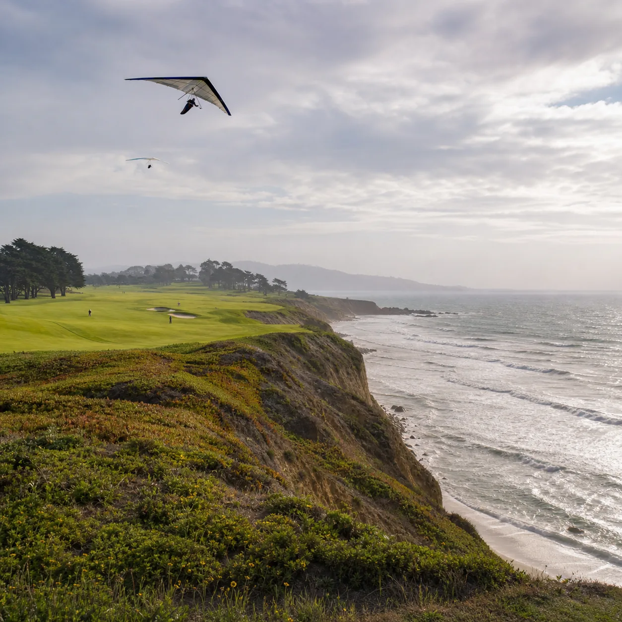 Realistic editorial photo of a coastal municipal golf course on high cliffs above the Pacific Ocean with hang glider sky