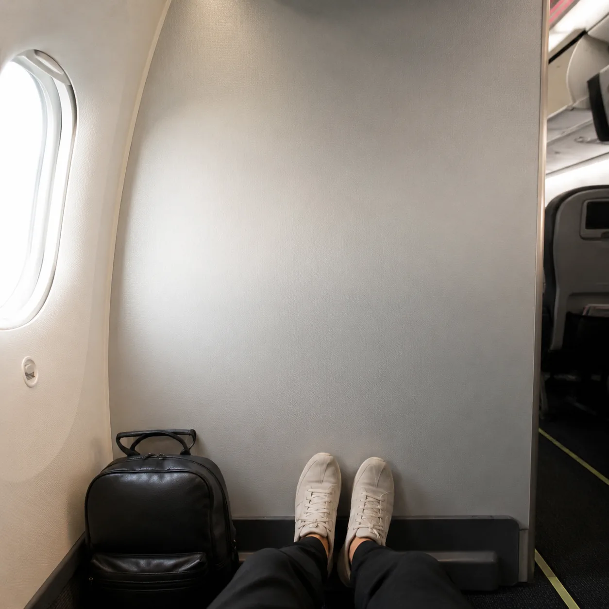 Realistic editorial photo of an airplane bulkhead row facing a solid cabin divider wall with a small personal item bag n