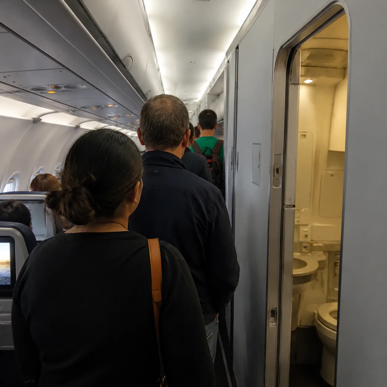 Realistic editorial photo of passengers standing in the aisle near an airplane lavatory during a long flight, realistic 