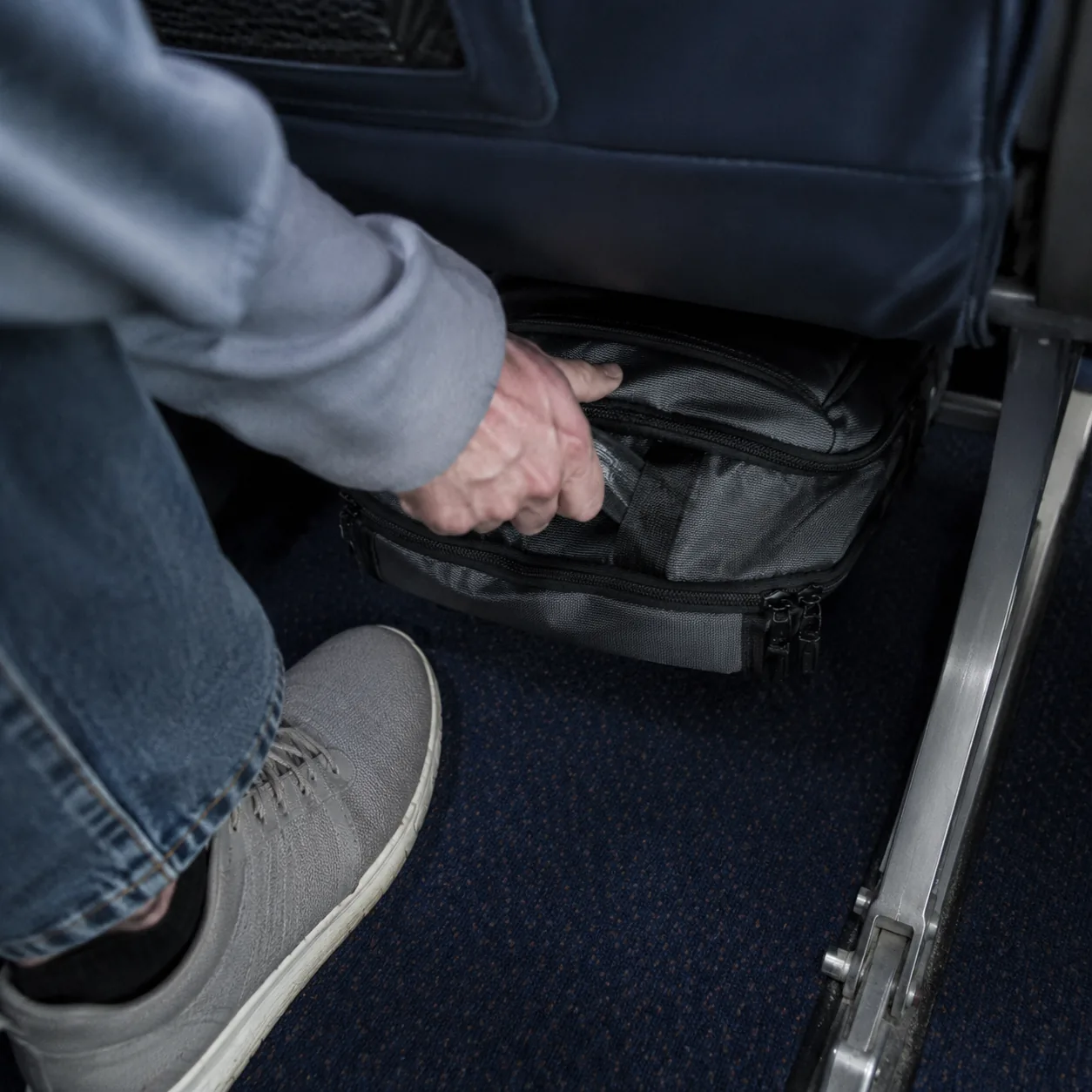Realistic editorial photo of a small backpack being placed under an airplane seat with limited foot space, realistic edi
