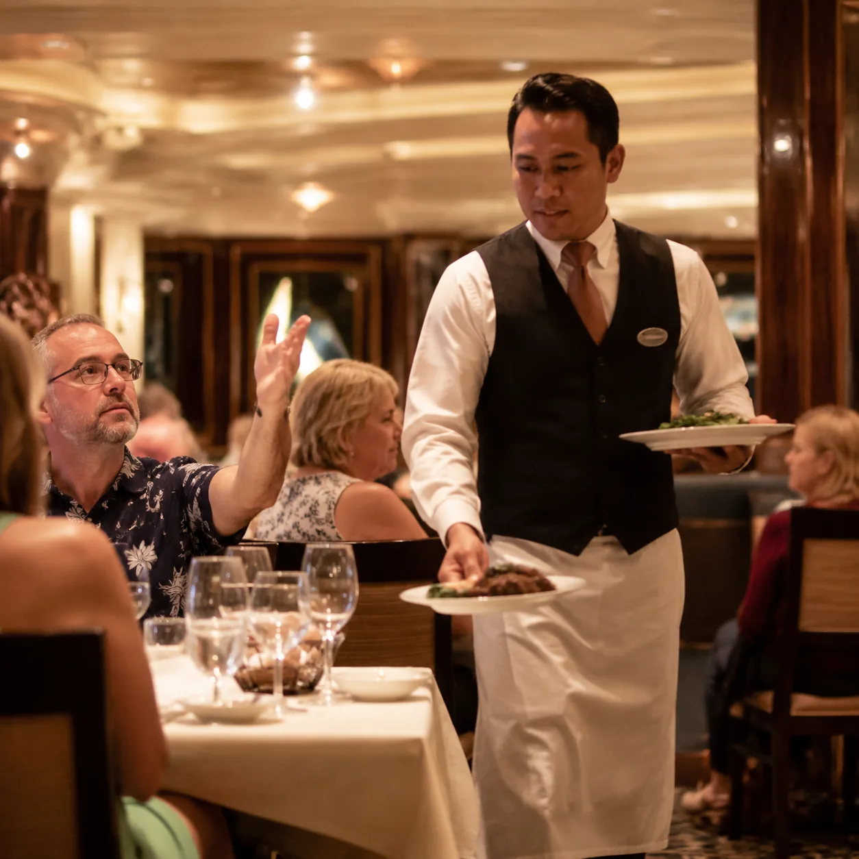 Realistic editorial photo of a cruise dining room server carrying plates while a passenger gestures impatiently from a t