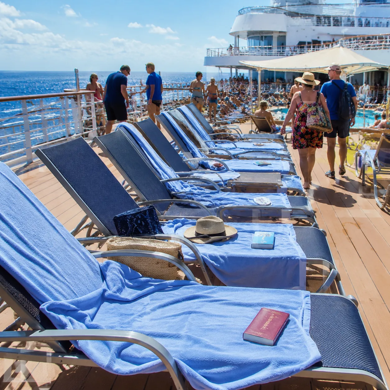 Realistic editorial photo of cruise ship pool deck loungers with towels and books reserving empty chairs while passenger