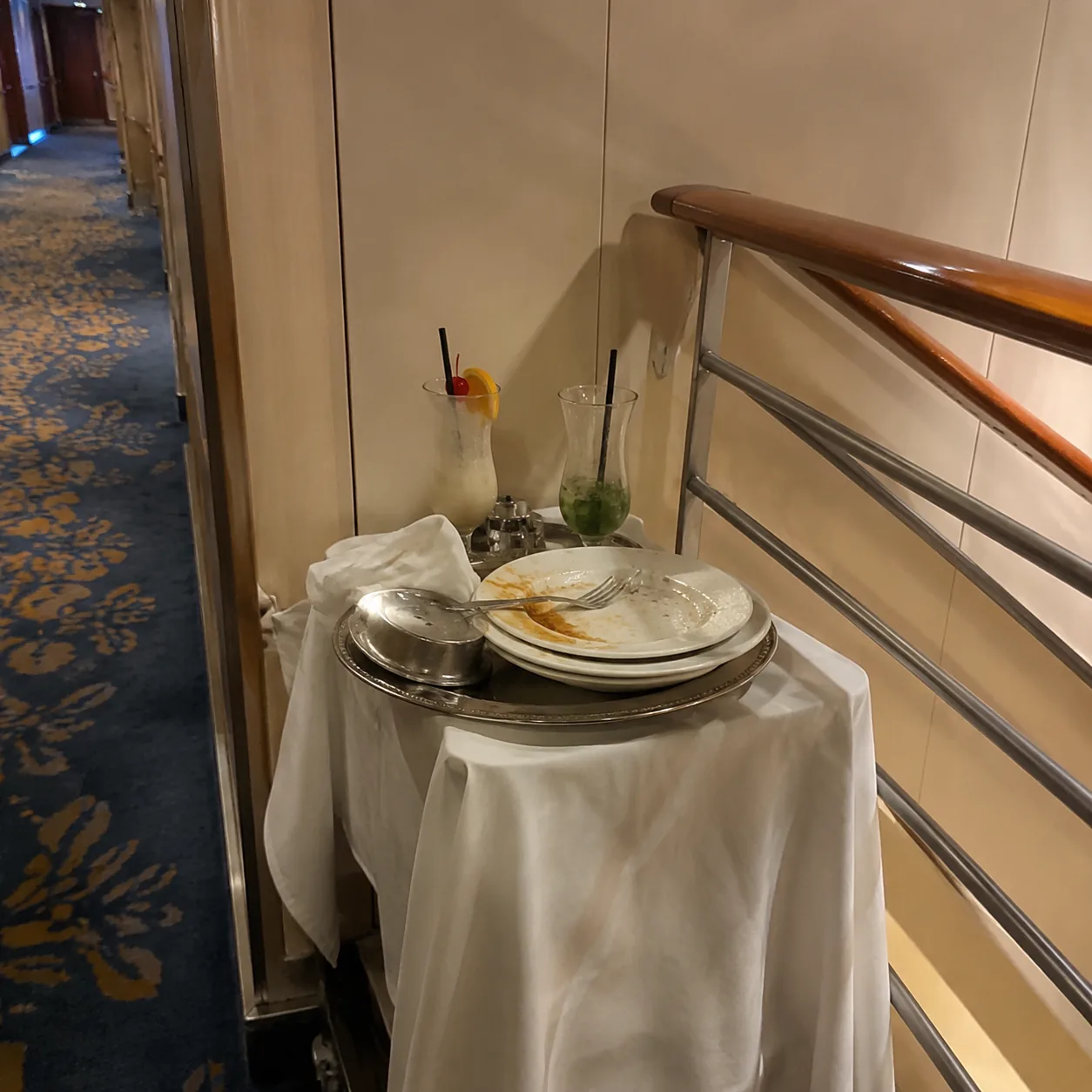 Realistic editorial photo of used cruise room service plates and cocktail glasses left beside a hallway wall and stairwe