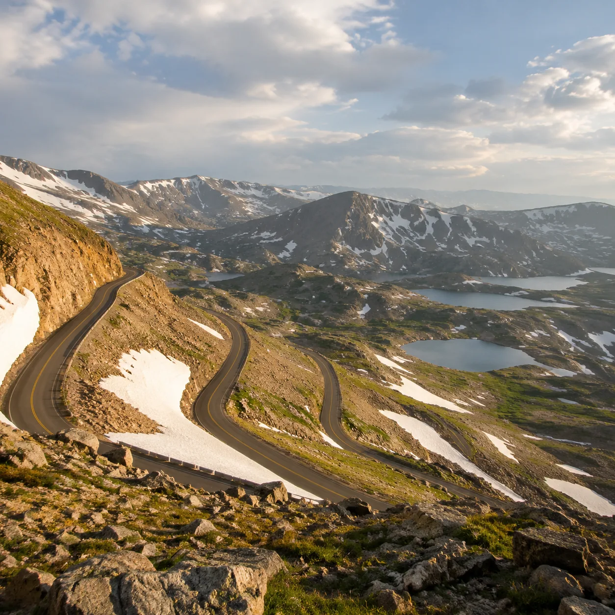 Realistic editorial photo of high alpine road and switchbacks on Beartooth Highway with snow patches and mountain lakes,