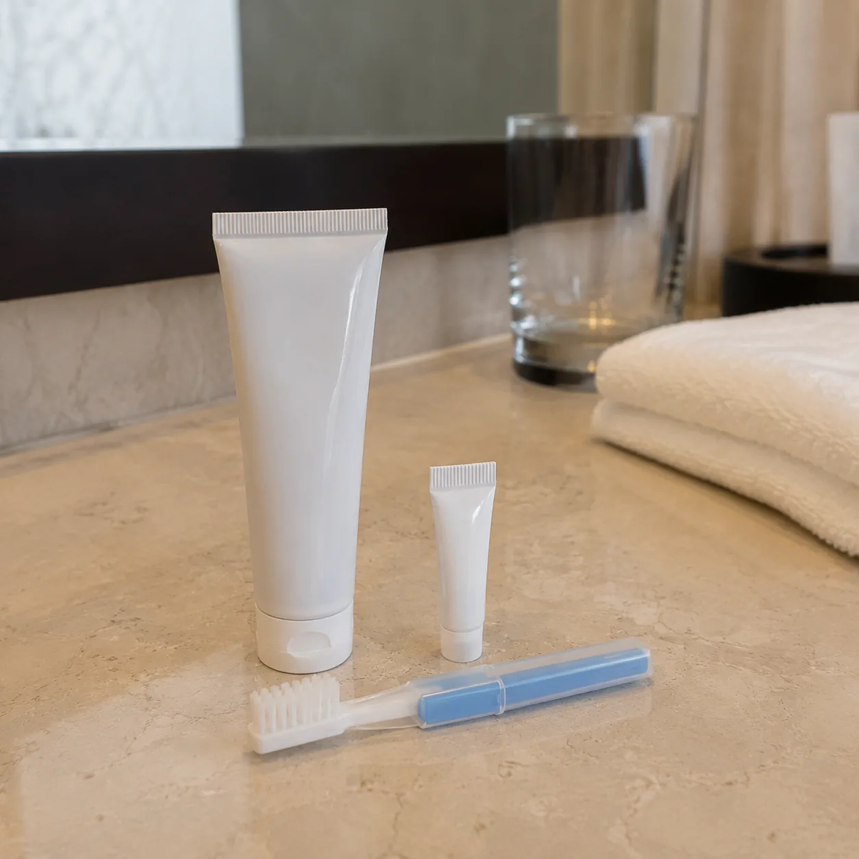 Realistic editorial photo of a standard toothpaste tube beside a tiny travel toothpaste and toothbrush on a hotel-style 