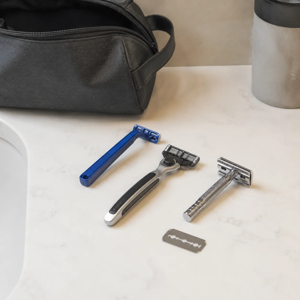Realistic editorial photo of disposable razor, cartridge razor, safety razor handle, and loose blade separated on a bath