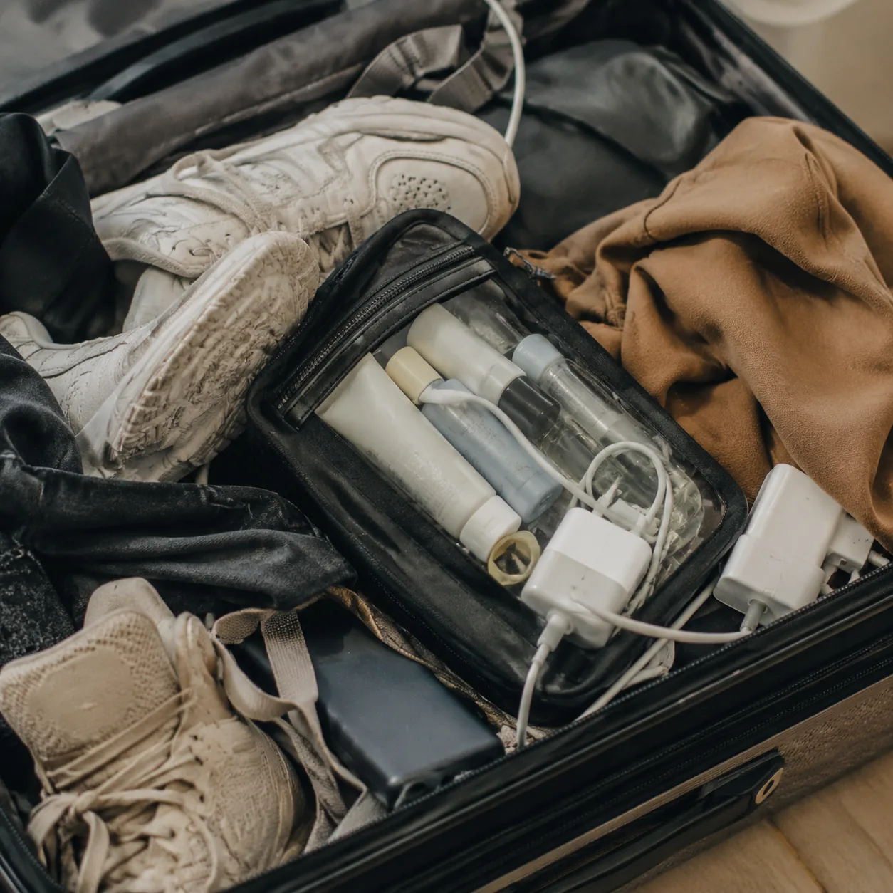 Realistic editorial photo of a clear quart toiletry bag buried under shoes, charging cables, and clothes inside an open 