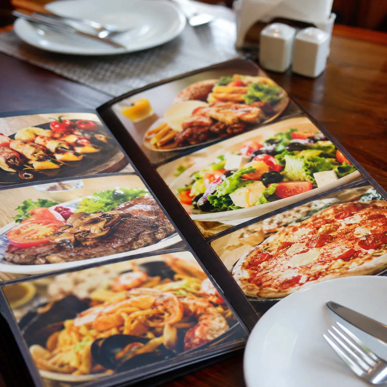 Realistic editorial photo of a glossy tourist restaurant menu with generic food photos on a table beside plates and cutl